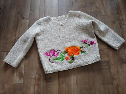 A white knitted sweater spread out on a dark wood laminate floor with a pattern of violet and orange flowers worked into its lower edge