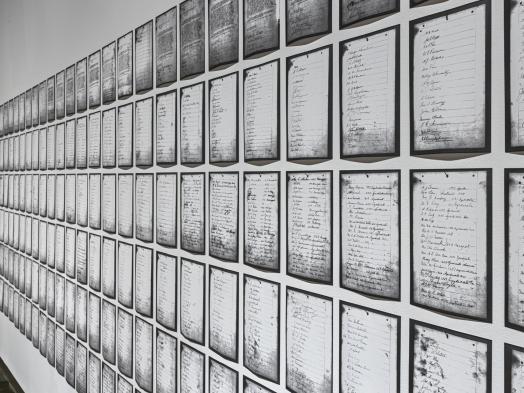 A sidelong view of Deanna Bowen's prints of a 1911 petition, showing black-edged letter-sized pages receding in rows along a white gallery wall