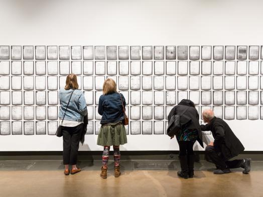 Photo of two pairs of gallery visitors seen from behind as they examine rows of pages of Deanna Bowen's installation of a 1911 petition, which fills the wall to both edges of the photo