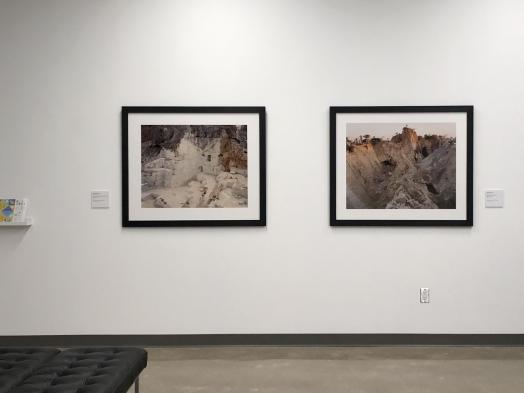 Installation view of two Edward Burtynsky photographs of marble quarries in black frames on a white lobby wall; the edge of a leather bench is visible in the lower left corner