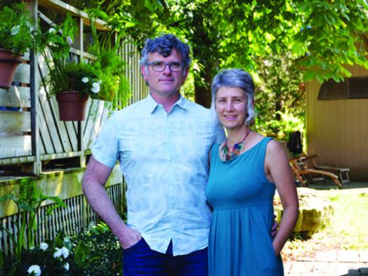 Photo of Trent Bauman and Juanita Metzger, an older couple casually dressed and standing together in a backyard filled with lush greenery