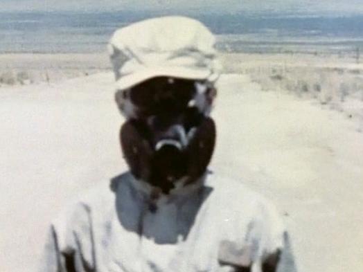 A film still from Trinity 3 shows a close-up view of a figure in light-coloured military fatigues and gas mask against a bleached desert landscape