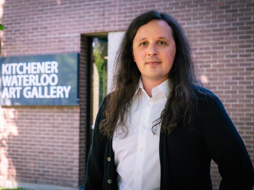 Photo of Darryn Doull, a young white man with long black hair wearing a white shirt and black cardigan, standing outside with the Kitchener-Waterloo Art Gallery's corner sign behind him