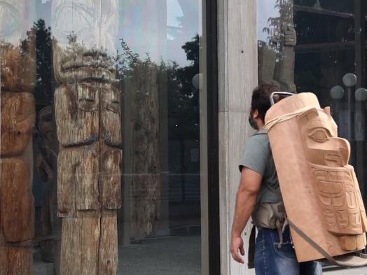 Video still of Luke Parnell's Remediation showing the artist wearing a Haida wood carving on his back, gazing at a totem pole displayed behind glass