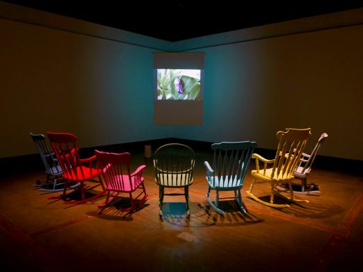 A semi-circle of colourful rocking chairs face a small video projection of a cocoon in a dark gallery