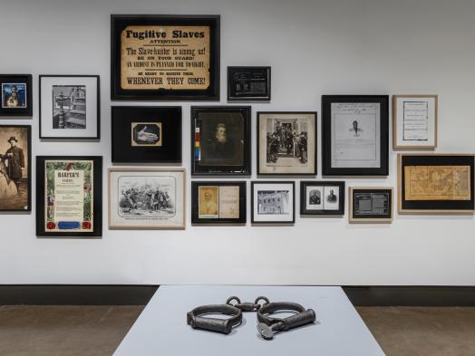 Installation view of Deanna Bowen's Black Drones in the Hive, showing a pair of shackles displayed on a white plinth in front of a wall filled with framed archival images