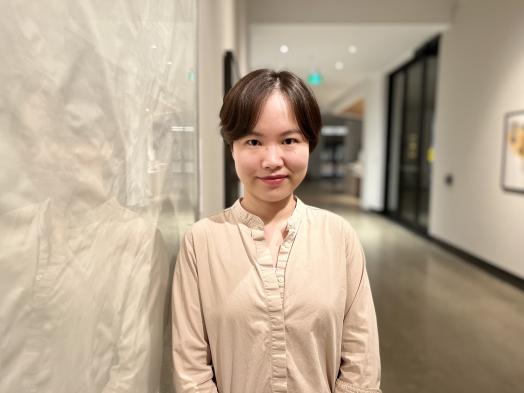 Photo of Yexin Tan, a young Chinese woman with short dark hair wearing a white shirt standing in the Corridor Gallery alongside a framed white Ron Martin work