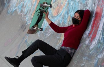 A female skateboarder in dark clothes and a black facemask holds up her green skateboard while reclined against the side of a graffiti-covered half-pipe wall