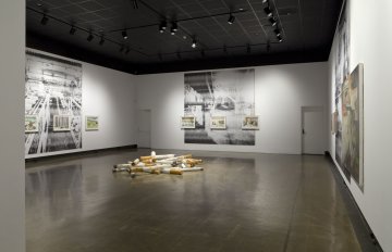 installation view of exhibition with large images on three walls and a large sculpture of cigarettes in the middle of the floor