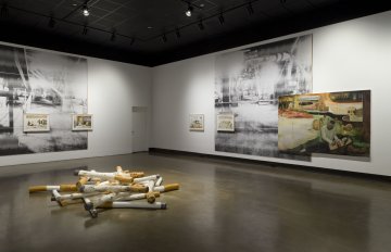 installation view of exhibition with large images on two walls and a large sculpture of cigarettes in the middle of the floor