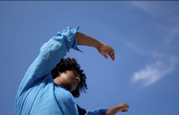 Black woman dancing in a blue shirt with the sky in the backdrop