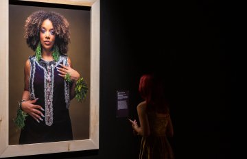 portrait of a black woman hung on a black wall being admired by a gallery visitor