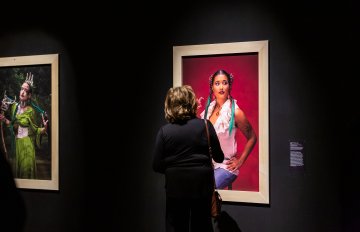 portrait of a women in a white translucent blouse hung on a black wall being admired by a gallery visitor