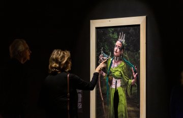 portrait of a woman in a green dress hung on a black wall being admired by a gallery visitor