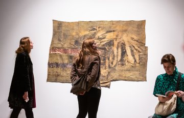 Beige fabric art piece hung on wall being admired by gallery visitors