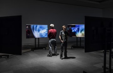 two people standing in the middle of a circular display of tv screens, all playing videos on them