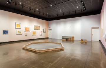 installation shot of gallery exhibition showcasing colourful pastel walls with frames hung on it and a hexagon shape made of wood in the centre
