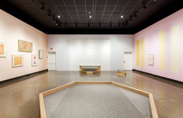 installation shot of gallery exhibition showcasing colourful pastel walls with frames hung on it and a hexagon shape made of wood in the centre