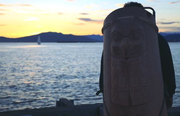 man with part of a totem pole strapped on his back looking out at a sunset on a lake