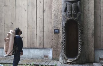 man looking at a totem pole while having part of a totem pole strapped to his back