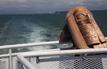 man with a totem pole on his back looking out at the water as he sails on a boat