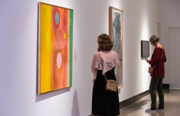 colourful abstract paintings hung on the wall being admired by gallery visitors