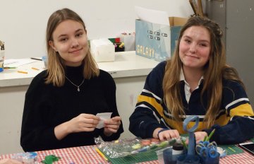 Photo of two female presenting youth smiling at the camera lens and crafting art using their hands.