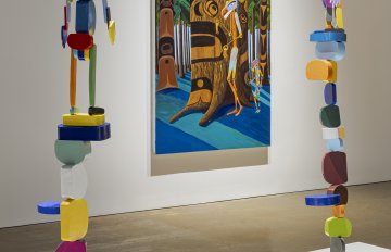 Two tall abstract ovoid figures presented on a white plinth in front of a painting