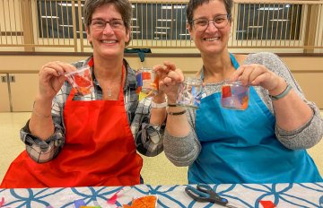 Two people wearing aprons sitting at a table smiling andholding up decoupage materials