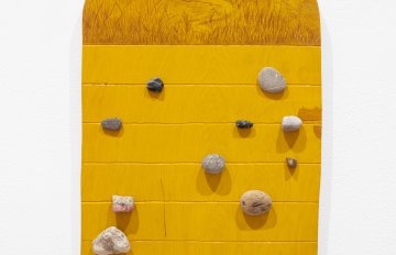 Yellow wooden board with a rounded top with brown pencil markings to depict a landscape around a body of water. Rocks are attached in random locations below