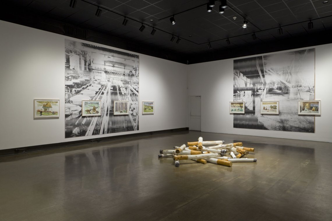 exhibition installation shot with large images on the walls and a large sculpture of a pile of cigarettes on the floor