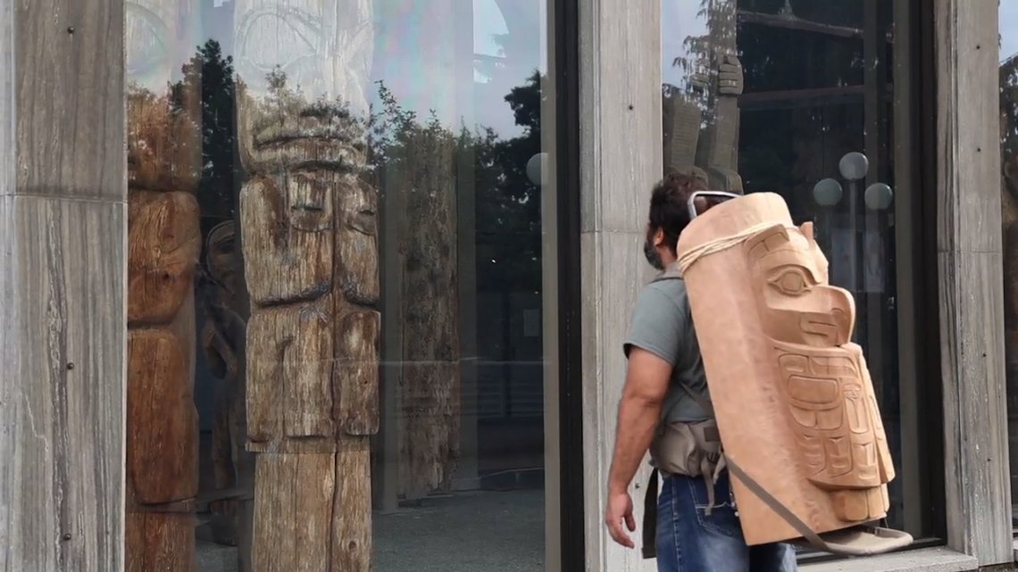 man looking in the window of a shop with a part of a toem pole on his back