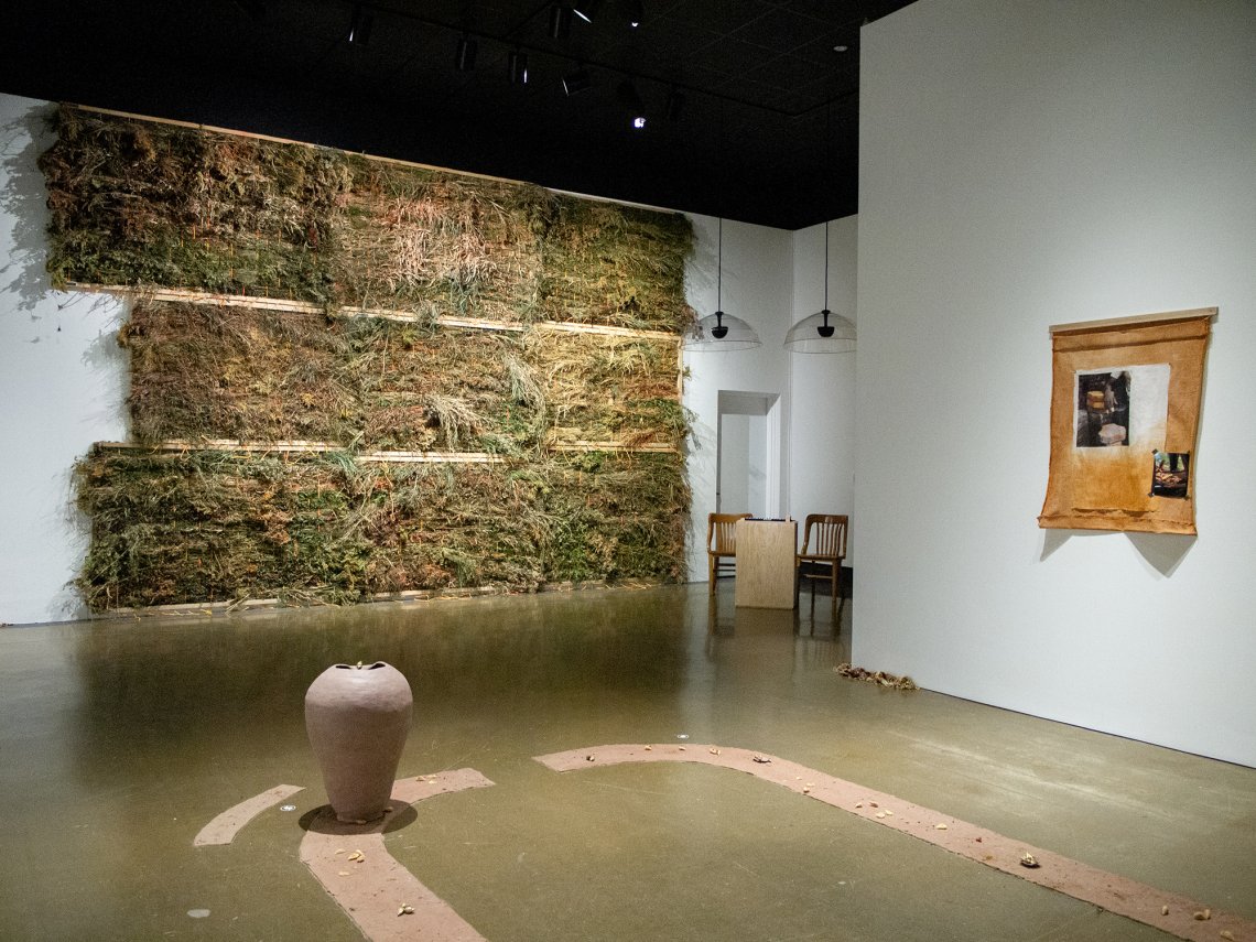 Gallery installation shot featuring a back wall of various dried plants, a hand made, unfired vessel and a piece of cloth with a photo on the right side wall