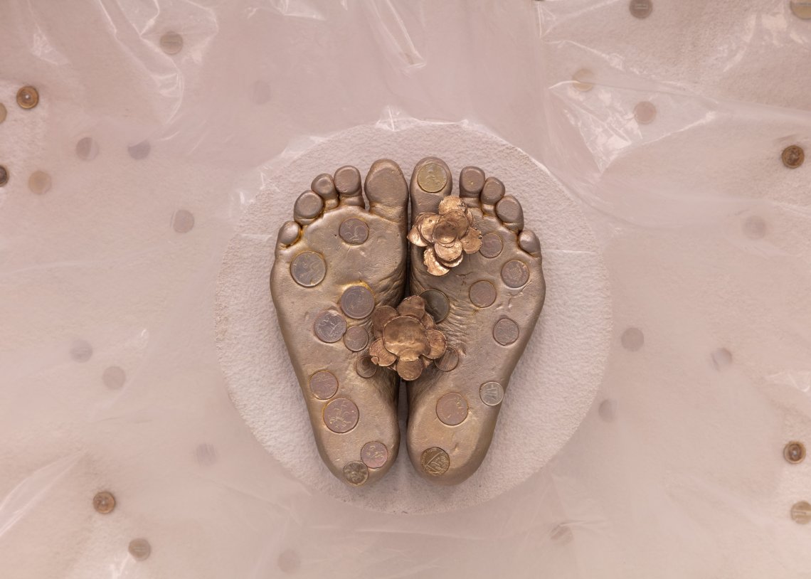 View of a casting of feet from underneath in gold on a background of concerete, interspersed with coins.