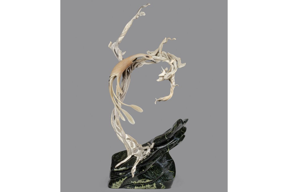 Photo of an antler and ivory sculpture standing on a base of labrodorite. The figure of a loon is leaping in the air to catch a fish.