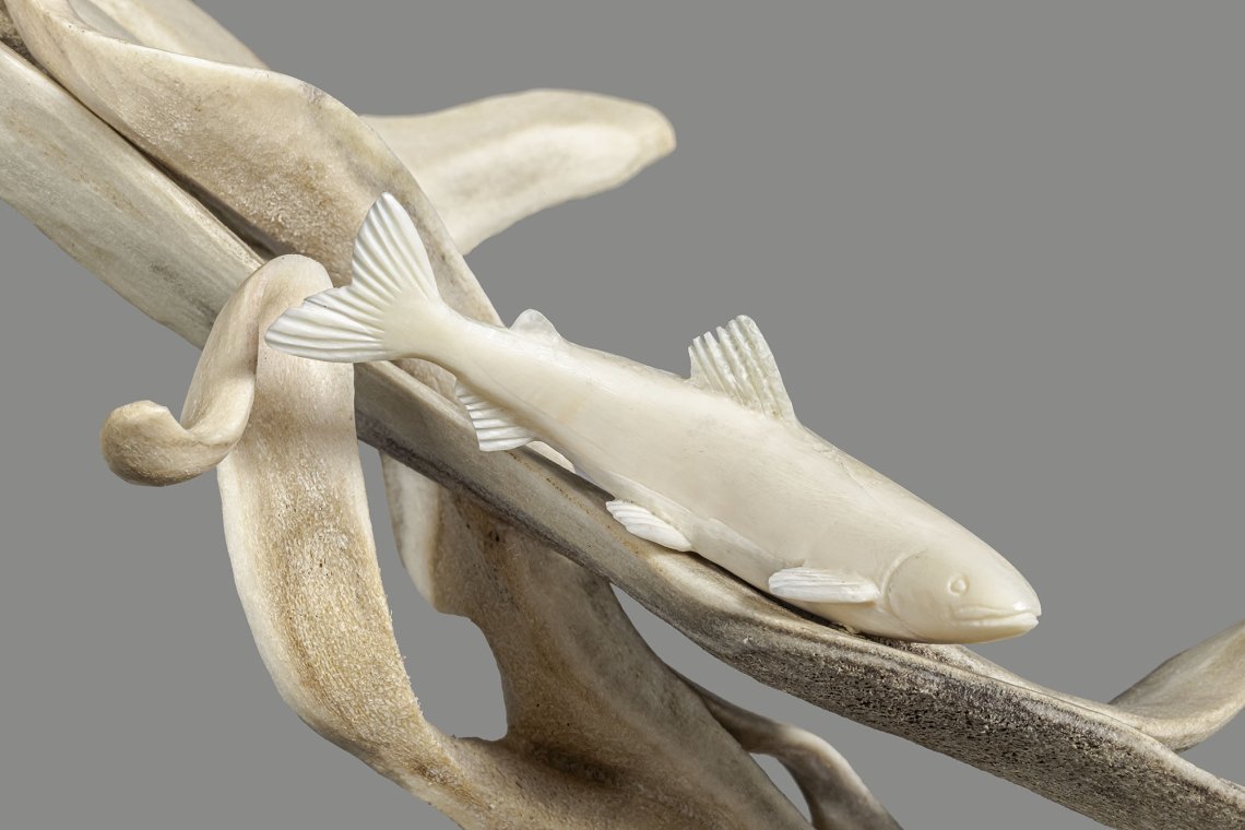 Closeup of a photo of an antler and ivory sculpture standing on a base of labrodorite. The figure of a loon is leaping in the air to catch a fish.