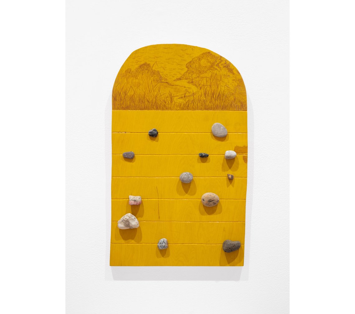 Yellow wooden board with a rounded top with brown pencil markings to depict a landscape around a body of water. Rocks are attached in random locations below