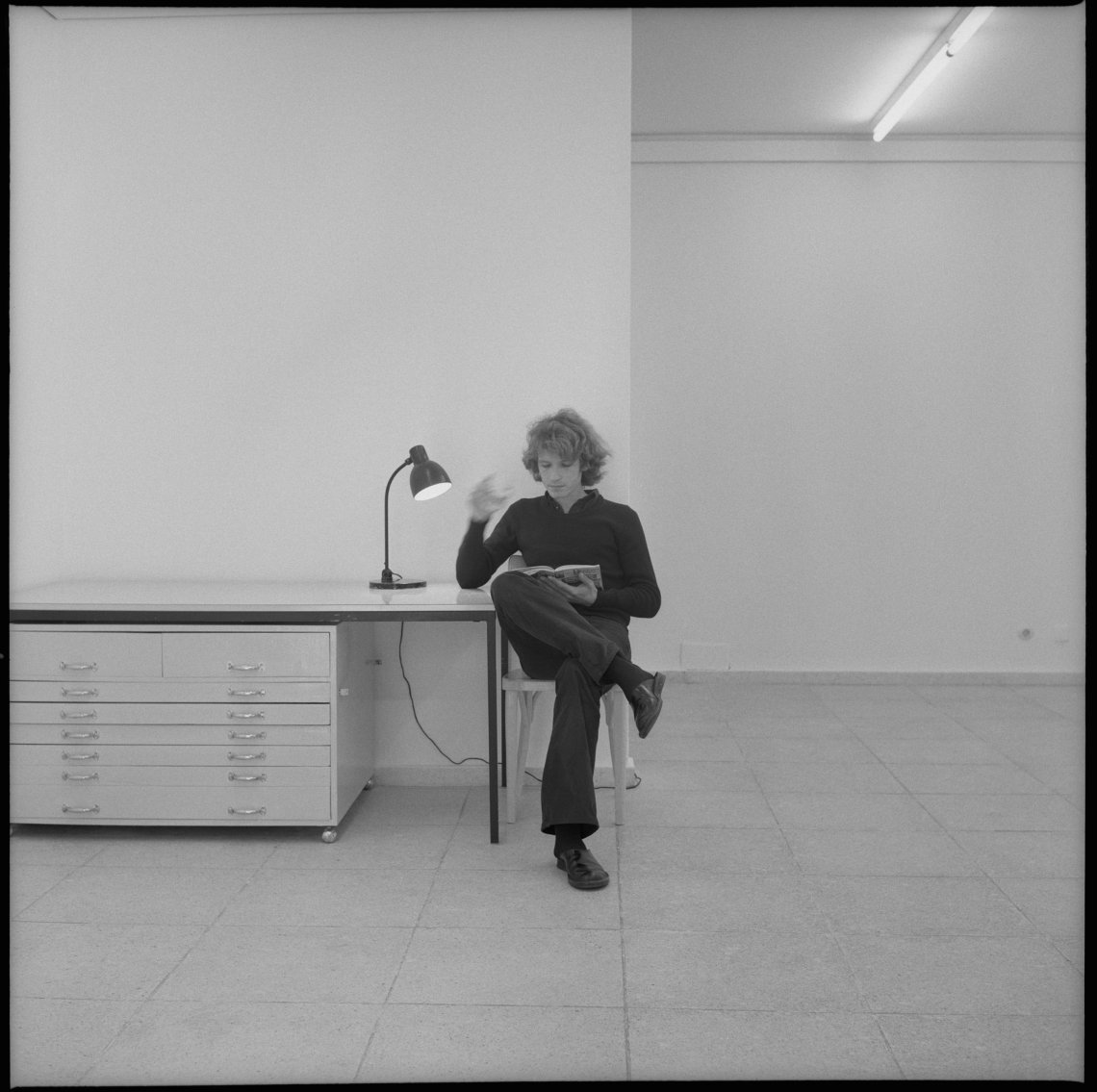 A man sitting on a chair reading a book and leaning on the table beside him.