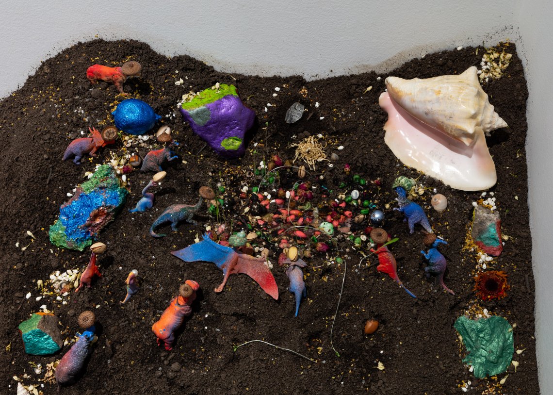 Mound of soil with colorful plastic dinosaurs and a seashell spread throughout with a small fairy door on the top side