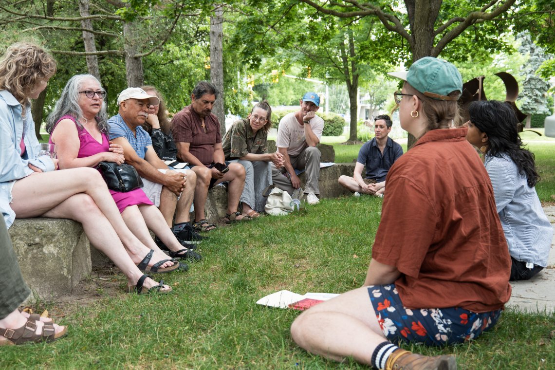 Photo of a group of individuals in a discussion. Some sit on the grassy floor, others sit on rocks.