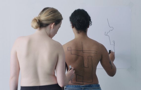 Image of the backs of two people. One person is drawing on the back of the person in front of them. The second person is tracing a line on the wall in front of them.