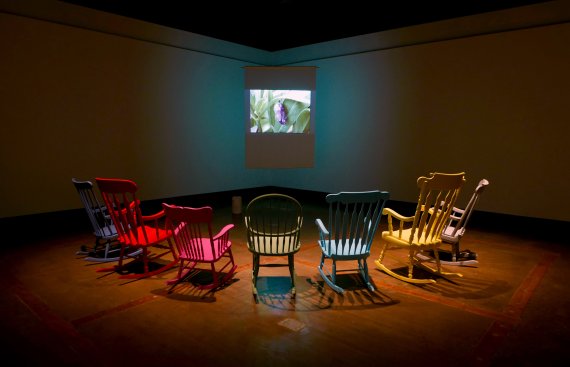 A semi-circle of colourful rocking chairs face a small video projection of a cocoon in a dark gallery