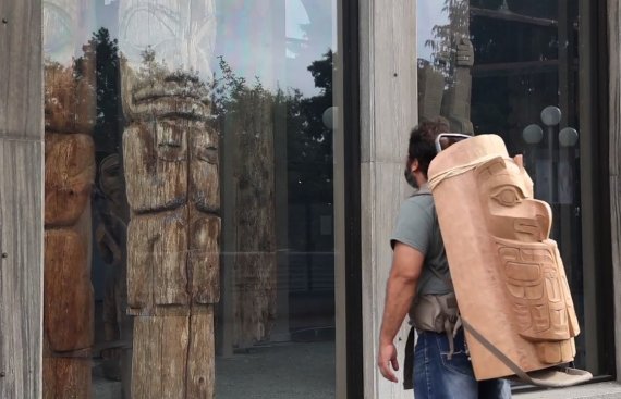 man looking in the window of a shop with a part of a toem pole on his back