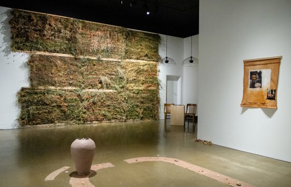 Gallery installation shot featuring a back wall of various dried plants, a hand made, unfired vessel and a piece of cloth with a photo on the right side wall