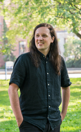 Photo of Darryn Doull, a young white man with long dark hair wearing a dark short-sleeved shirt, standing with hands in pockets in an outdoor park setting