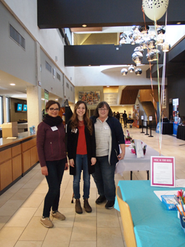 A photo of three women with volunteer nametags standing together and smiling next to a table prepared with art supplies in a theatre lobby with balloons in the background
